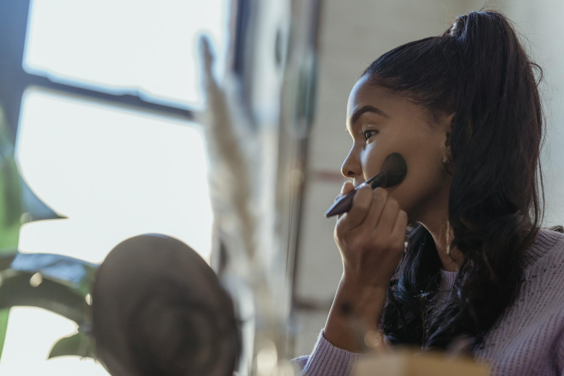 A young woman confidently applies makeup indoors, using a brush near a window.