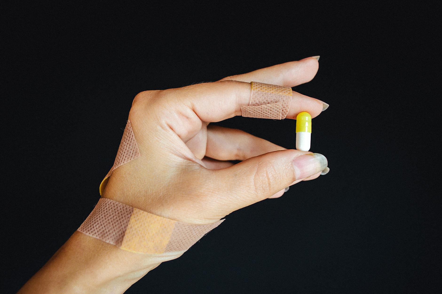 A hand with bandages holding a yellow and white capsule against a black background.