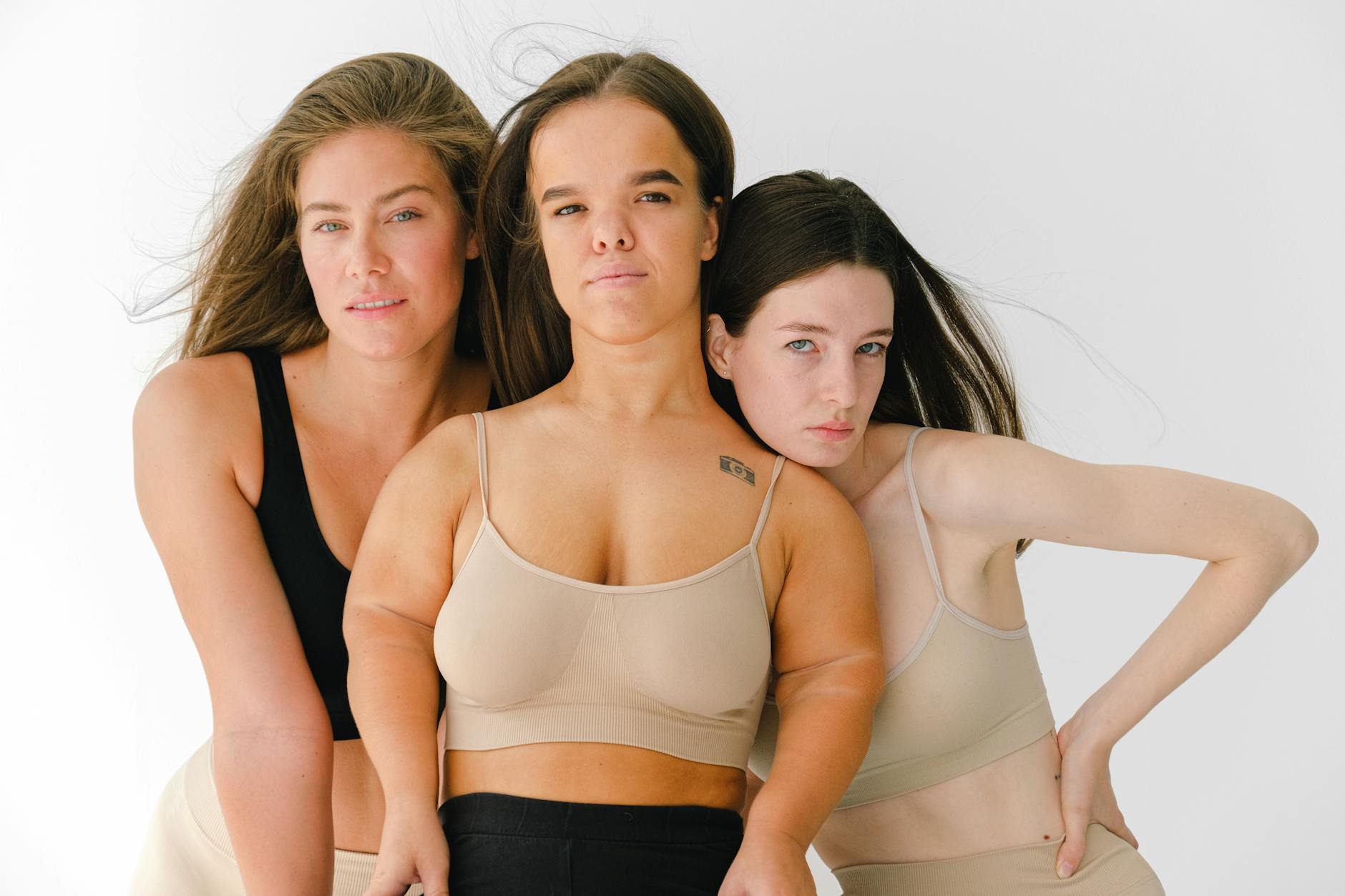 Three women confidently posing in sports bras, embodying body positivity and diversity in a studio setting.